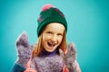 Portrait of cheerful child girl wears winter mittens, warm sweater, hat with pompom and openwork cape scarf on blue