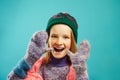 Portrait of cheerful child girl wears winter mittens, warm sweater, hat with pompom and openwork cape scarf on blue