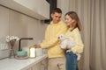 Portrait of cheerful couple in the kitchen, man washing the dishes in the sink, woman holding little white dog in hand, Royalty Free Stock Photo