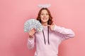 Portrait of cheerful curly haired teenage girl in hoodie with halo on head pointing finger on bunch of dollar banknotes and