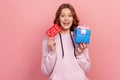 Portrait of cheerful curly haired teenage girl in hoodie holding sale word and wrapped gift box, low price on presents, discount Royalty Free Stock Photo