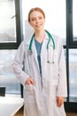 Portrait of cheerful female doctor in white coat standing on background of window in sunny day in light medical clinic Royalty Free Stock Photo