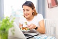 Cheerful female who is working with laptop at the table at the home and eating salad