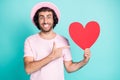 Portrait of cheerful hipster guy pointing paper heart wear pink cap t-shirt spectacles isolated on teal color background