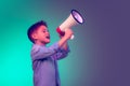 Portrait of cheerful little boy, child shouting in megaphone, posing isolated over green purple background in neon light Royalty Free Stock Photo