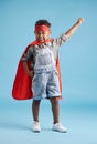 Portrait of cheerful little boy in superhero cape and mask stretching out his hand on blue background. Strong brave kid