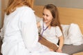 Portrait of cheerful little girl listening heartbeat with stethoscope to female doctor sitting in bed. Royalty Free Stock Photo