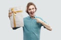 Portrait of cheerful modern young man in light blue shirt standing and pointing finger to present with yellow bow and toothy smile Royalty Free Stock Photo