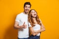 Portrait of cheerful people man and woman in basic clothing smiling and showing thumb up at camera, isolated over yellow