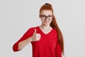 Portrait of cheerful redhaired female with freckled skin, shows like, raises thumb, approves somebody`s actions, dressed in red cl