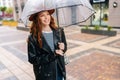 Portrait of cheerful redhead young woman wearing fashion hat standing with transparent umbrella on city street near Royalty Free Stock Photo