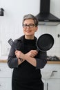 Portrait of a cheerful smiling mature woman with a frying pan on the background of the kitchen Royalty Free Stock Photo