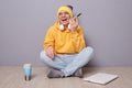 Portrait of cheerful smiling positive man wearing beanie hat, yellow hoodie and jeans posing against gray wall, sitting on floor Royalty Free Stock Photo