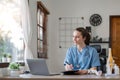 Portrait of cheerful smiling young female doctor in blue medical uniform typing on laptop computer, sitting at desk near Royalty Free Stock Photo