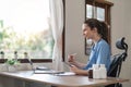 Portrait of cheerful smiling young female doctor in blue medical uniform typing on laptop computer, sitting at desk near Royalty Free Stock Photo