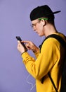 Portrait of a cheerful student wearing backpack, in cap and glasses and using smartphone over purple background Royalty Free Stock Photo