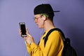Portrait of a cheerful student wearing backpack, in cap and glasses and using smartphone over purple background Royalty Free Stock Photo
