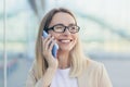 Portrait of a cheerful woman with glasses blonde holding a mobile phone smiling talking on a smartphone near the office Royalty Free Stock Photo