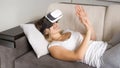 Portrait of young cheerful woman lying with VR headset on sofa and trying to catch something Royalty Free Stock Photo