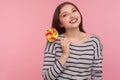 Portrait of cheerful woman in striped sweatshirt holding sweet round rainbow candy lollipop, pink background