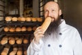 Portrait of cheerful young adult baker with long beard in white uniform standing in his manufacture, smelling with pleasure fresh Royalty Free Stock Photo