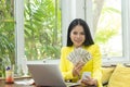 Portrait of cheerful young Asian woman holding money banknotes and mobile phone with smile face in front of computer notebook Royalty Free Stock Photo