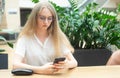 Portrait of a cheerful young caucasian blond business woman sitting on the table, holding and looking at phone and thinking. On Royalty Free Stock Photo
