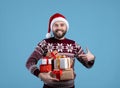 Portrait of cheerful young man in warm sweater and Santa hat pointing at heap of gifts on blue studio background