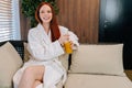 Portrait of cheerful young woman wearing white bathrobe enjoying drinking cocktail sitting on sofa by window at spa Royalty Free Stock Photo