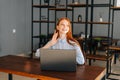 Portrait of cherrful young redhead woman sitting at desk with laptop in modern office room and looking away. Royalty Free Stock Photo