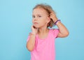 Portrait of cherubic blue-eyed little girl titivating touching short curly fair hair, wearing pink jumpsuit, bracelets.