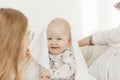 Portrait of cherubic smiling happy blue-eyed plump baby infant toddler covered with white blanket sitting on bed linen.