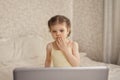 Girl 3 - 4 years Caucasian in yellow dress sits in the bedroom of bright bed and looking at laptop covering his mouth in Royalty Free Stock Photo