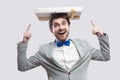 Portrait of childish funny young man in gray coat and blue bow tie standing and pointing fingers to present on his head, toothy