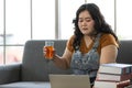 Portrait of chubby asian woman wearing casual clothing sitting on a couch holding a glass of herbal juice looking at a laptop