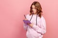 Portrait of clever curly haired teenage girl in hoodie writing in notebook, completing to-do list or studying Royalty Free Stock Photo