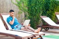 Portrait of clever handsome bearded young adult freelancer man in blue t-shirt and shorts lying on cozy deck chair with laptop on