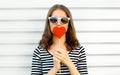 Portrait close-up woman kissing red heart shaped lollipop or hides her lips on white wall