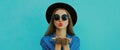 Portrait close up young woman blowing her red lips sending sweet air kiss wearing a black round hat, denim jacket on a blue Royalty Free Stock Photo