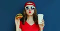 Portrait close up of young woman with burger and coffee cup wearing baseball cap, sunglasses over blue Royalty Free Stock Photo