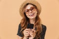 Portrait closeup of content woman wearing straw hat and sunglasses laughing and holding smartphone, isolated over beige background Royalty Free Stock Photo