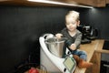 Portrait of concentrated little boy toddler with fair hair sitting on table near kneader cooking machine in kitchen.