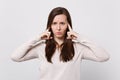 Portrait of concerned young woman in light clothes looking camera covering ears with fingers on white wall