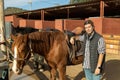 Portrait of experienced adult stables worker in plaid shirt standing close to saddled horse in countryside club Royalty Free Stock Photo