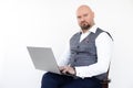 Portrait of confident bald businessman in grey vest, blue jeans, white shirt, sitting on chair, holding laptop, posing. Royalty Free Stock Photo