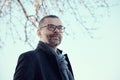 Portrait of a confident bearded middle-aged man with glasses wearing a blue coat and woollen scarf against the sky.