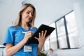 Portrait of confident female doctor standing in Hospital corridor. Beautiful nurse wearing blue scrubs, holding Royalty Free Stock Photo