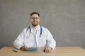 Portrait of confident joyful and smiling male doctor sitting at table on gray concrete background.