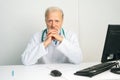 Portrait of confident mature adult male doctor in white coat sitting at desk with computer in medical office, looking at Royalty Free Stock Photo