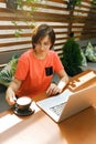 Portrait of confident mature professional woman in glasses, a coral T-shirt sitting on summer terrace in cafe, using laptop Royalty Free Stock Photo